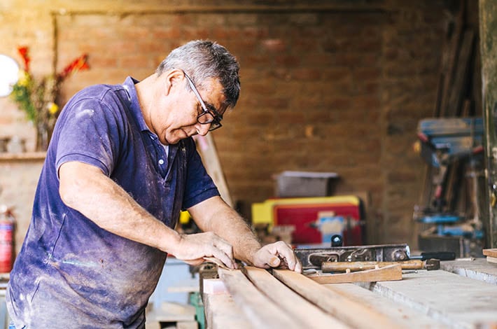 an older man wearing protective eyewear stands at a work shop sanding and cutting wood