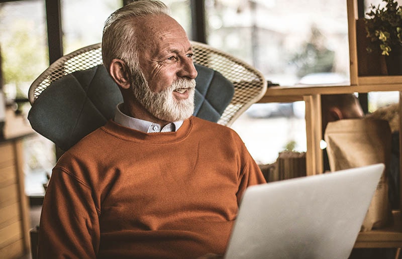 Man with white hair and a white beard in an orange sweater sits in a chair with a laptop in his lap