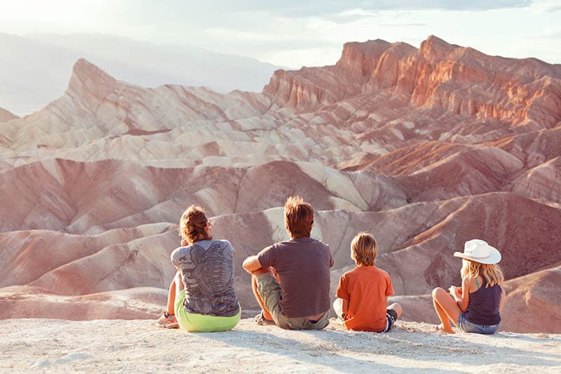 A family with 2 kids sit on the ground looking out at mountain scenery
