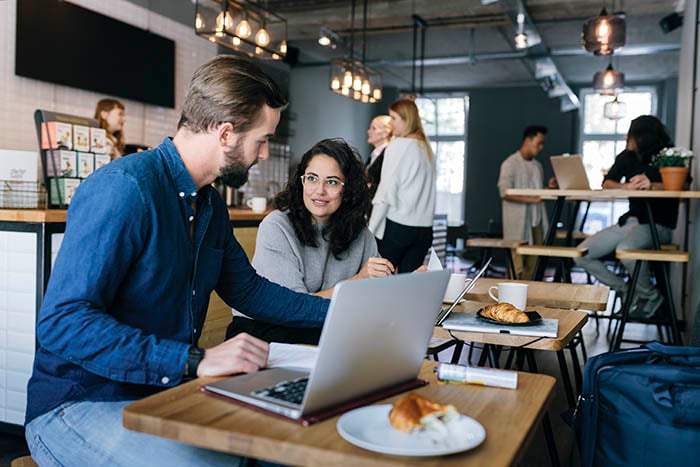 a man and a woman sit together at a coffee shop working together on laptops