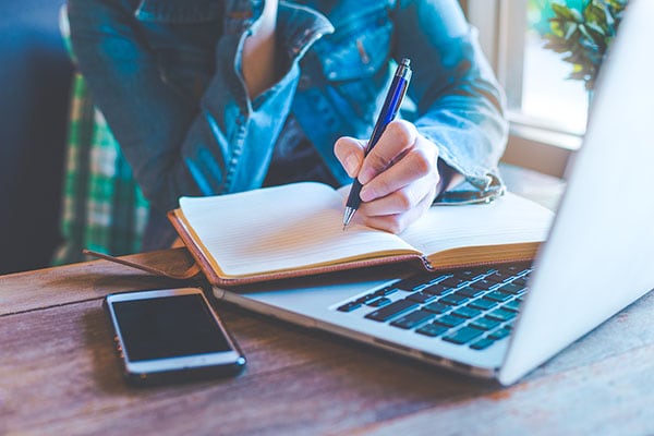woman in a denim shirt writes in a notebook with her laptop and her cell phone nearby on a wooden desk