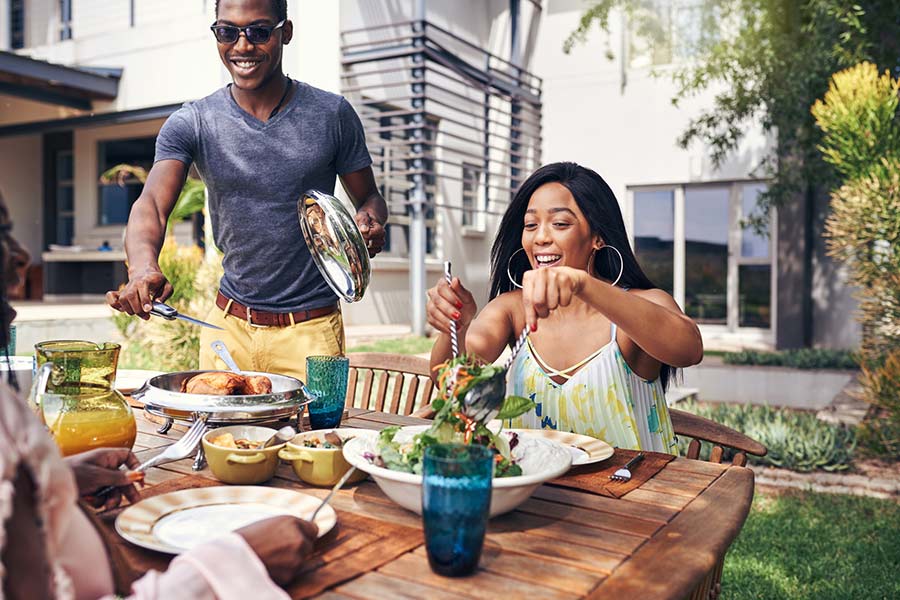 At a table in a house backyard, a woman tosses a salad while man prepares to cut chicken.