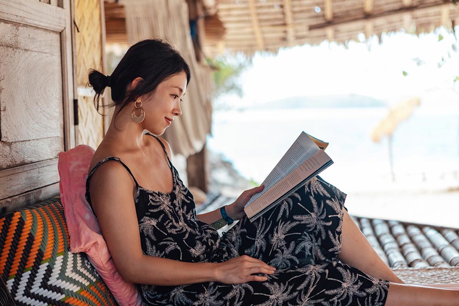A woman wearing a black palm tree dress reads a book in a cabana by the beach.