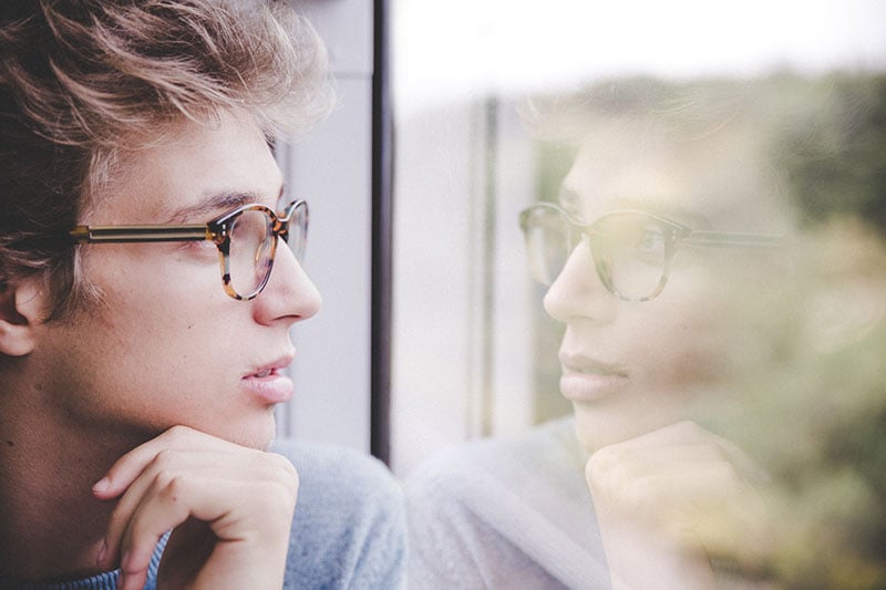 Young adult man wearing glasses looks out the window at some trees
