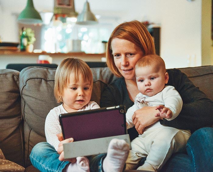 Red haired woman with a red haired toddler and a red haired baby, sitting on a gray couch and looking at an tablet