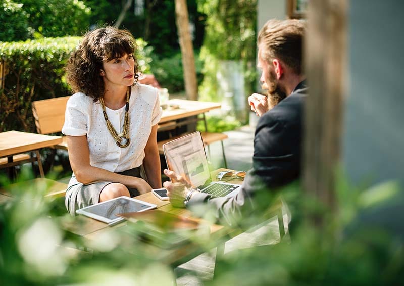 a view of a woman and a man at a table with a laptop in an outdoor cafe as they discuss something serious