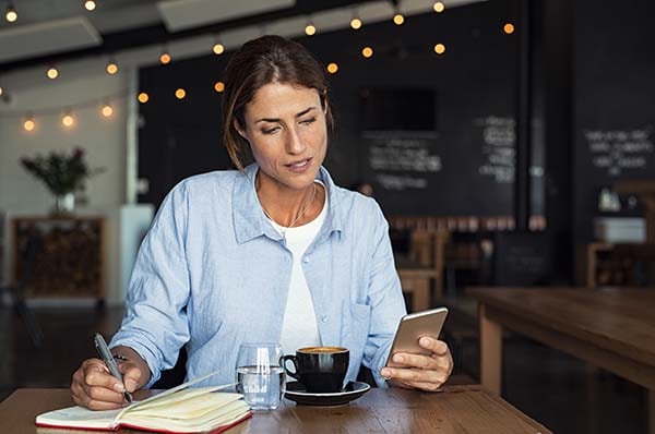 woman taking notes as she refers to her mobile device