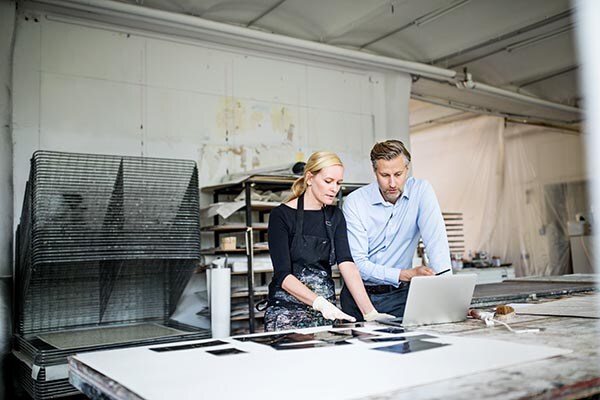 a woman and a man stand together over a work table while looking at a laptop