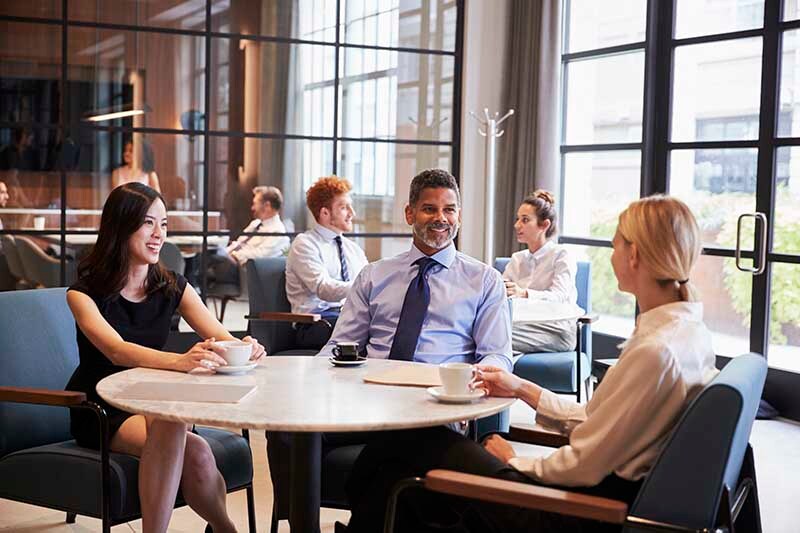 Two women and a man sit at a table smiling and talking. they are in a shared coworking space with 2 other people in the background.