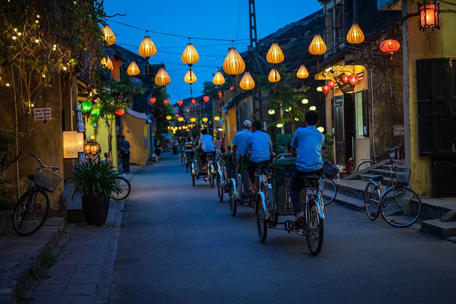 A group of bicycle cabs pedal down a dark street with lanterns along sides of the buildings